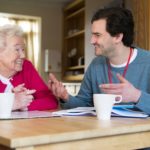 A horizontal shot of a male care worker sitting chatting with a senior woman, they are discussing some paperwork and smiling at one another while drinking cups of tea.